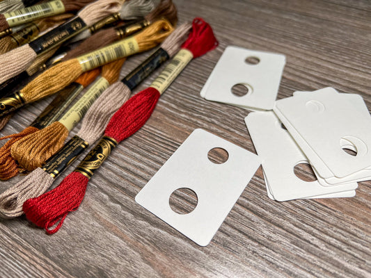 a pile of DMC embroidery floss skeins sitting next to a pile of blank hanging floss holders