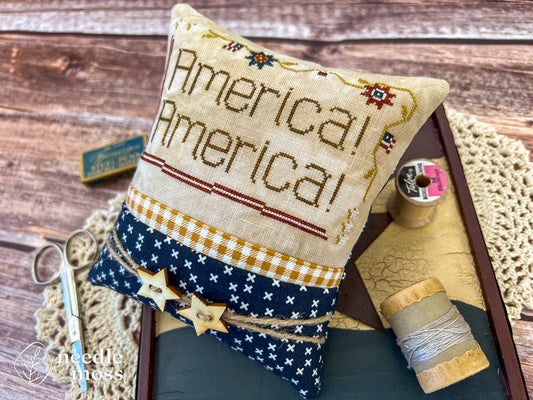 American patriotic cross stitch pillow with "America! America!" text and flag motifs sitting on a wood table surrounded by antique decorations and sewing notions