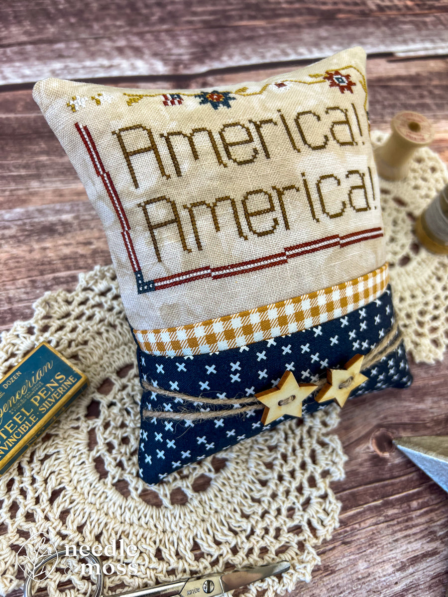 American patriotic cross stitch pillow with "America! America!" text and flag motifs sitting on a wood table surrounded by antique decorations and sewing notions