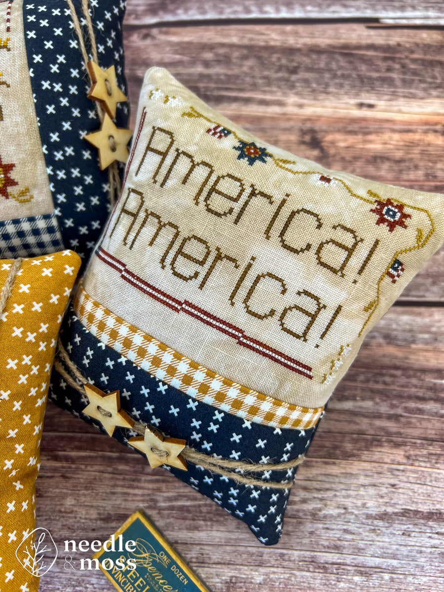 American patriotic cross stitch pillow with "America! America!" text and flag motifs sitting on a wood table surrounded by antique decorations and sewing notions