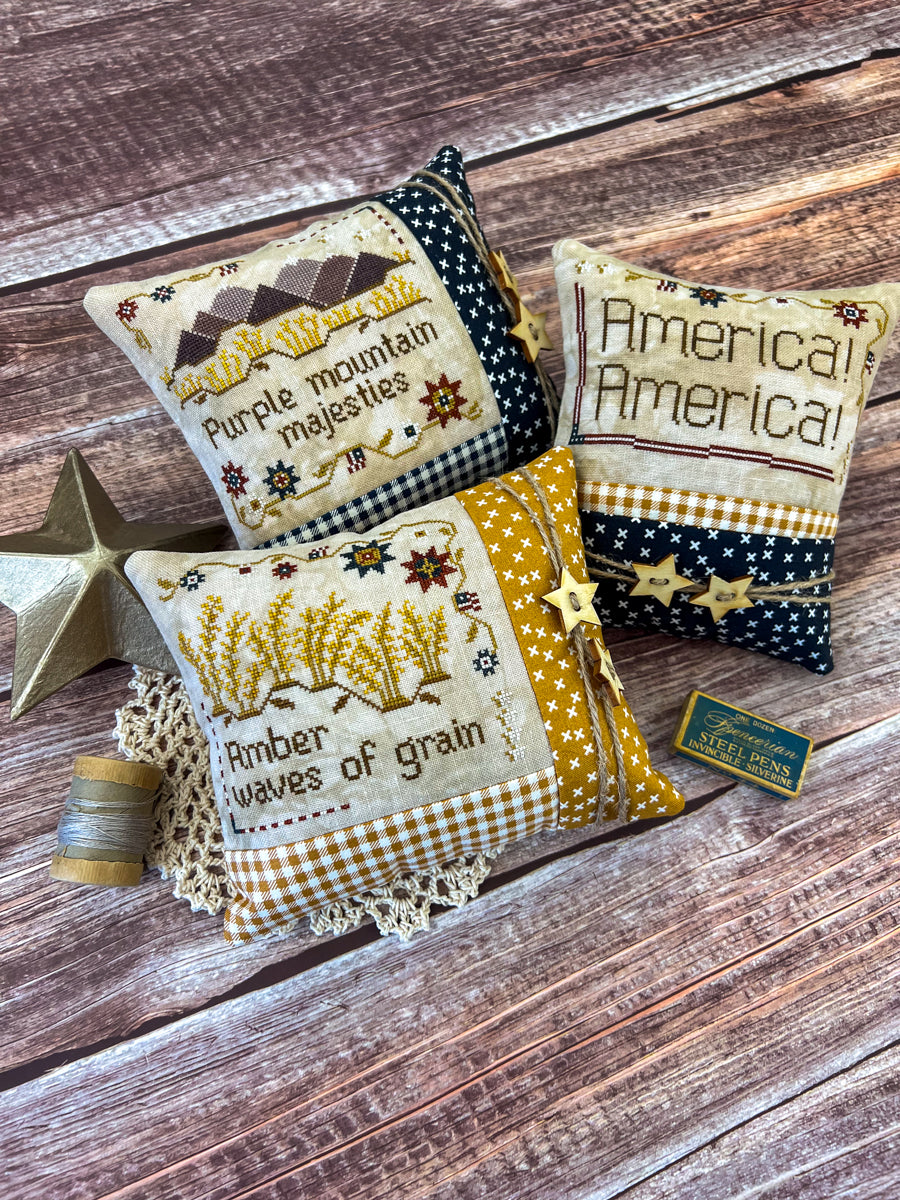 Three American patriotic pillows featuring words from the song "America the Beautiful" sitting on a wood table next to antique decorations and sewing notions