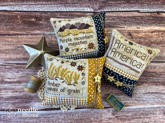 Three American patriotic pillows featuring words from the song "America the Beautiful" sitting on a wood table next to antique decorations and sewing notions