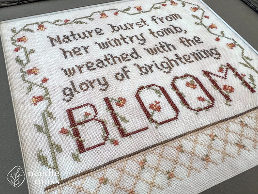 Close up view of a framed floral cross stitch piece that says "Nature burst from her wintry tomb wreathed with the glory of brightening bloom" stitched on light pink fabric with a dark gray mat surrounding it