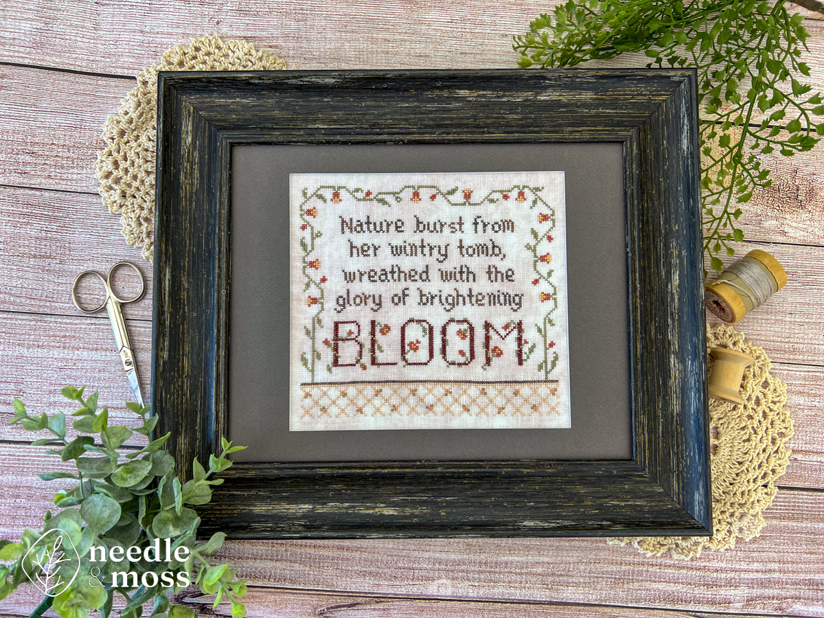 A framed floral cross stitch piece that says "Nature burst from her wintry tomb wreathed with the glory of brightening bloom" laying on a wooden table surrounded by sewing notions and leafy plants