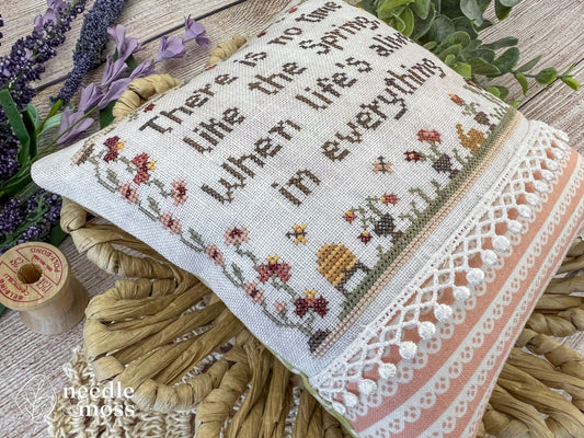 Close up view of a cross stich pillow showing spring flowers and motifs with white lace trim and pink finishing fabric sitting in a basket on a bed of flowers