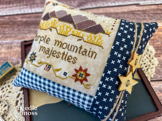 American patriotic cross stitch pillow with "Purple mountain majesties" text and mountain motifs sitting on a wood table surrounded by antique decorations and sewing notions