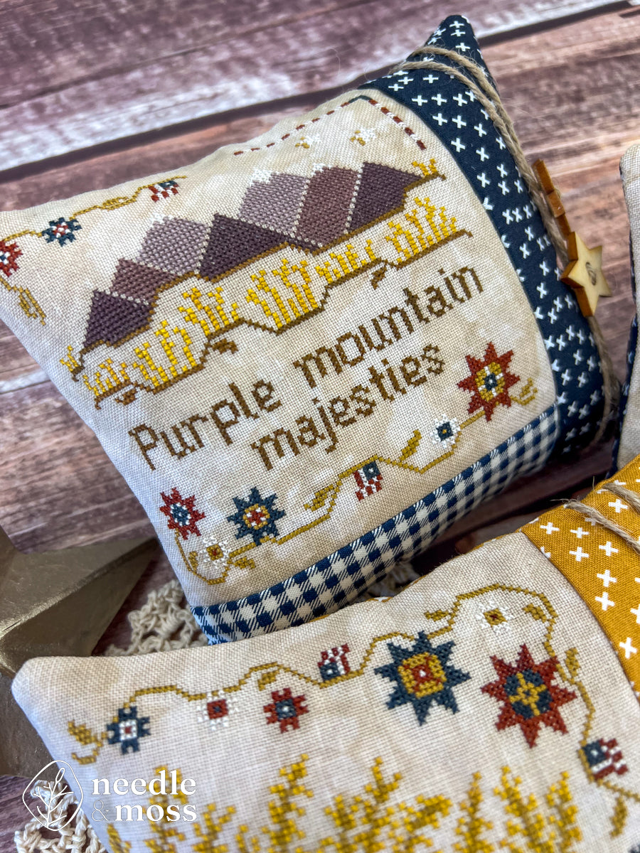 American patriotic cross stitch pillow with "Purple mountain majesties" text and mountain motifs sitting on a wood table surrounded by antique decorations and sewing notions