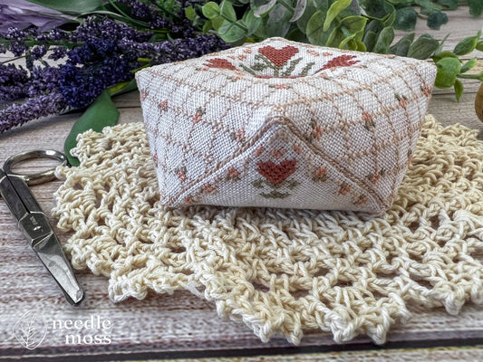 A handmade biscornu pinkeep with a quilted cross stitch pattern and pink heart flowers sitting on a crochet doily next to antique scissors surrounded by flowers