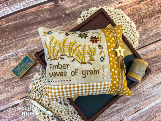 American patriotic cross stitch pillow with "Amber waves of grain" text and grassy field motifs sitting on a wood table surrounded by antique decorations and sewing notions