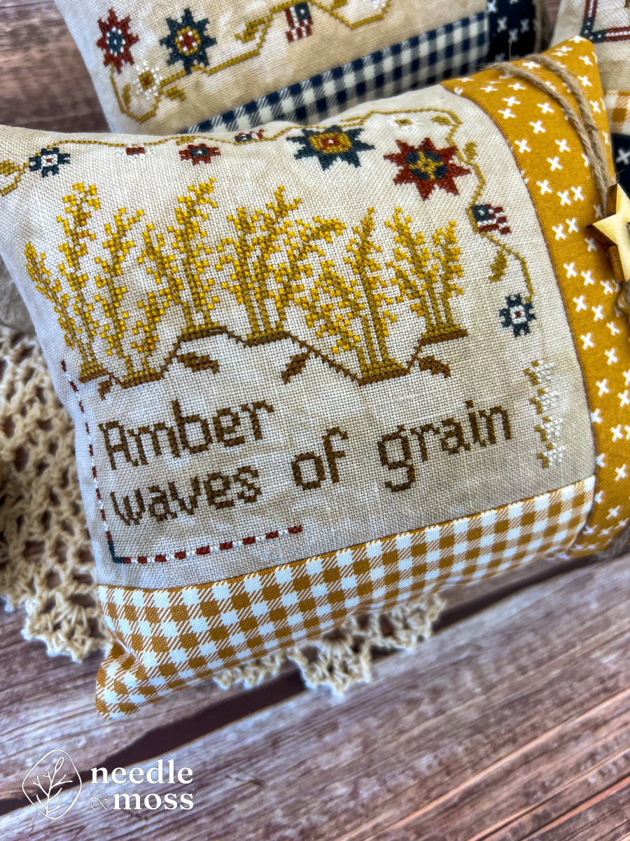 American patriotic cross stitch pillow with "Amber waves of grain" text and grassy field motifs sitting on a wood table surrounded by antique decorations and sewing notions