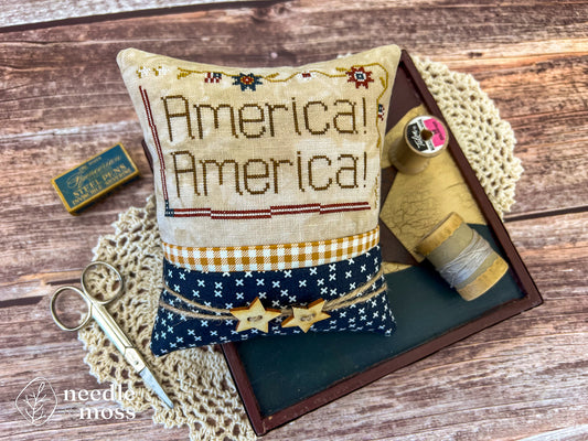 American patriotic cross stitch pillow with "America! America!" text and flag motifs sitting on a wood table surrounded by antique decorations and sewing notions