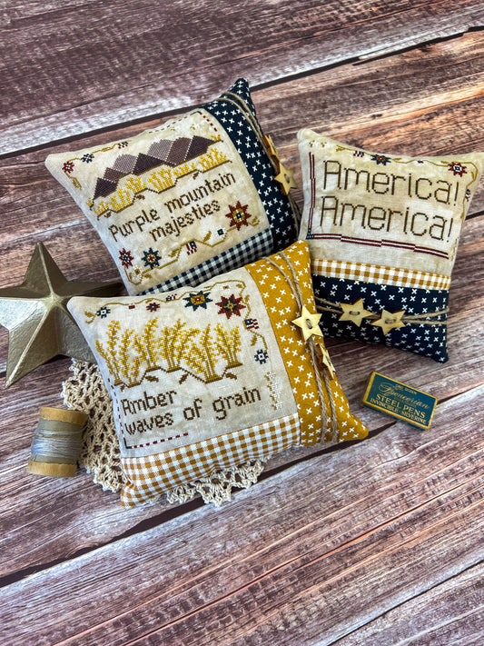 Three American patriotic pillows featuring words from the song "America the Beautiful" sitting on a wood table next to antique decorations and sewing notions