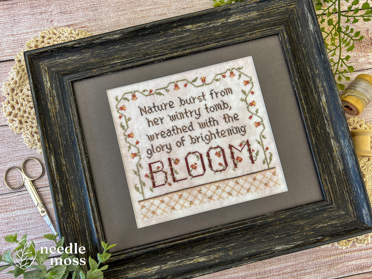 A framed floral cross stitch piece that says "Nature burst from her wintry tomb wreathed with the glory of brightening bloom" laying on a wooden table surrounded by sewing notions and leafy plants