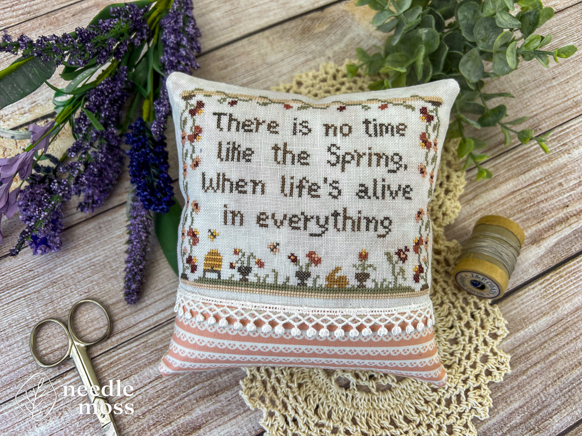 A floral spring cross stitch pillow that says "There is no time like the spring, when life's alive in everything" laying on a wood table surrounded by sewing notions and flowers