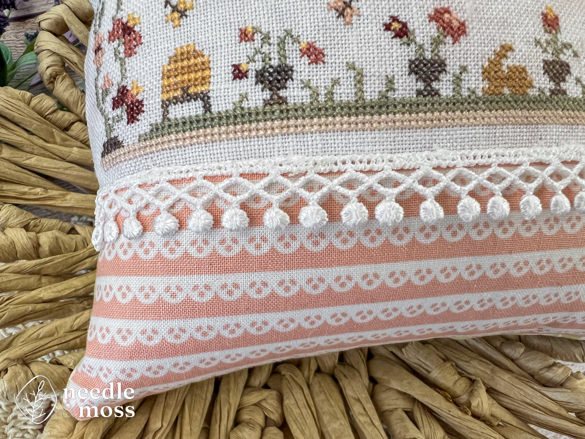 Close up view of a cross stich pillow showing spring flowers and motifs with white lace trim and pink finishing fabric sitting in a basket on a bed of flowers