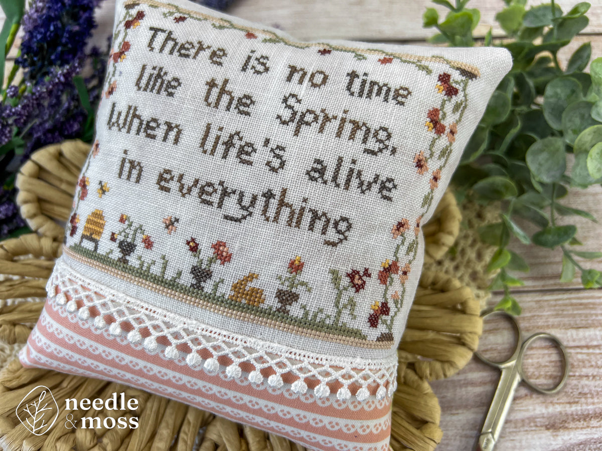 A floral spring cross stitch pillow that says "There is no time like the spring, when life's alive in everything" laying on a wood table surrounded by sewing notions and flowers