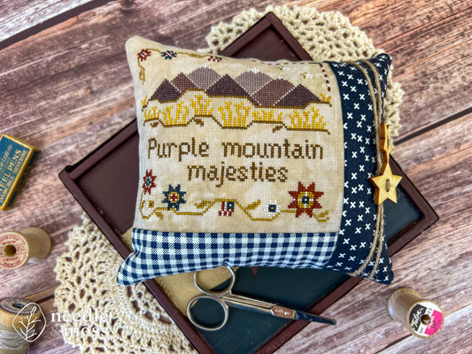 American patriotic cross stitch pillow with "Purple mountain majesties" text and mountain motifs sitting on a wood table surrounded by antique decorations and sewing notions