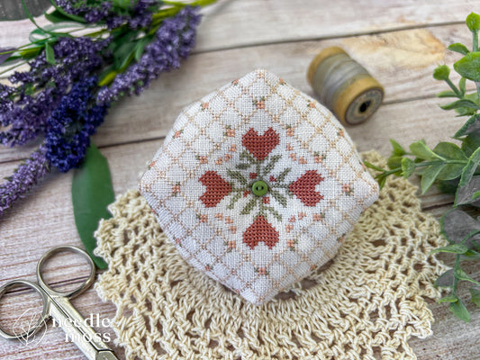 A handmade biscornu pinkeep with a quilted cross stitch pattern and pink heart flowers sitting on a crochet doily next to antique scissors and thread, surrounded by flowers