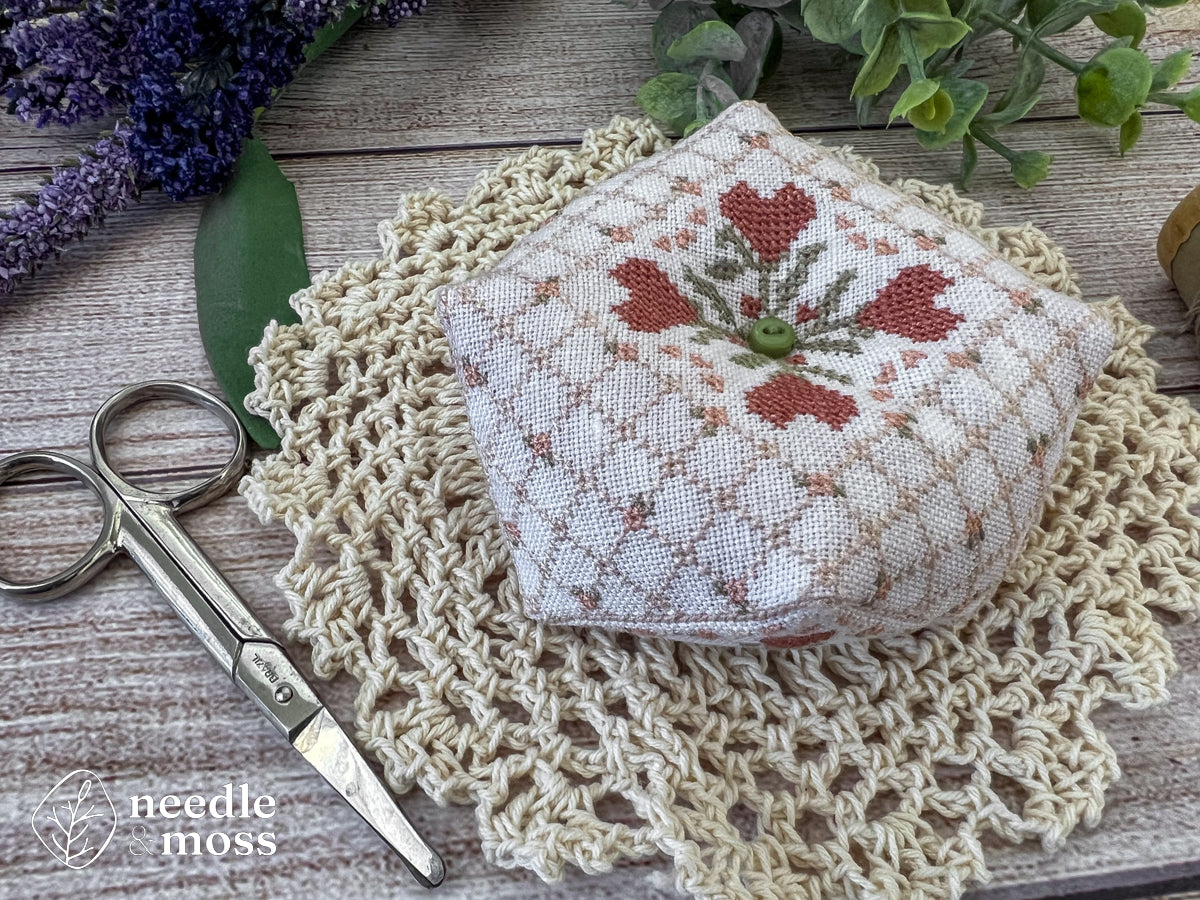 A handmade biscornu pinkeep with a quilted cross stitch pattern and pink heart flowers sitting on a crochet doily next to antique scissors surrounded by flowers